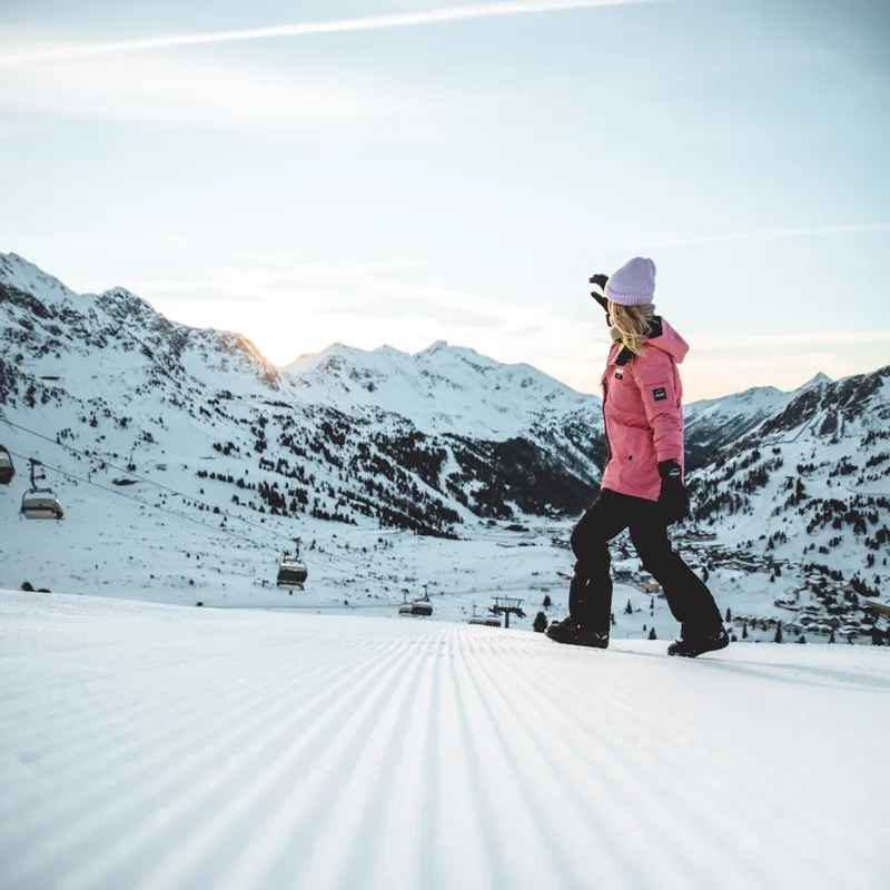Woman in pink ski jacket walking on groomed slope with snowy mountain background at sunrise